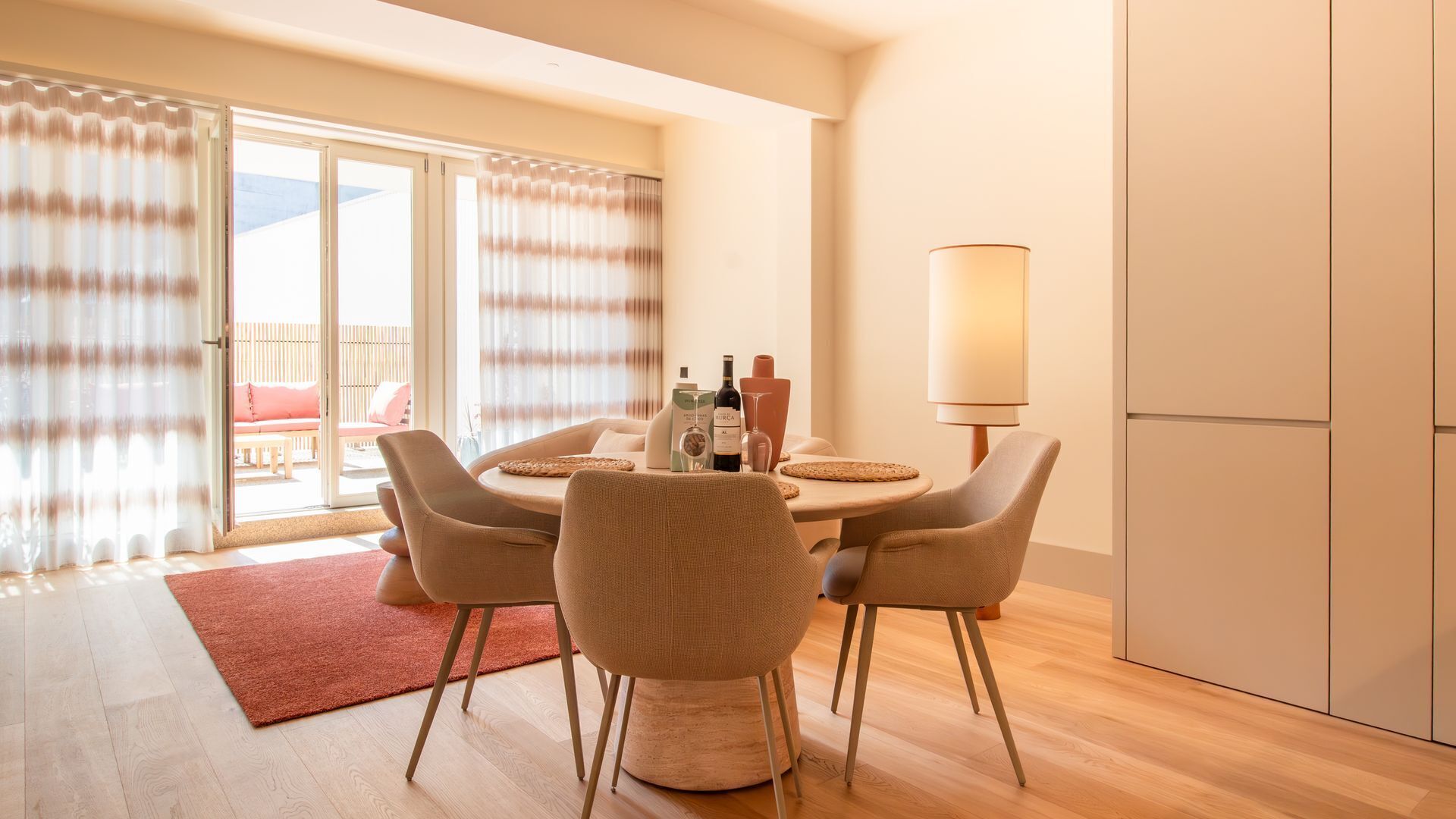 A dining area with a round table, chairs, a lamp, and a view of the outside balcony.
