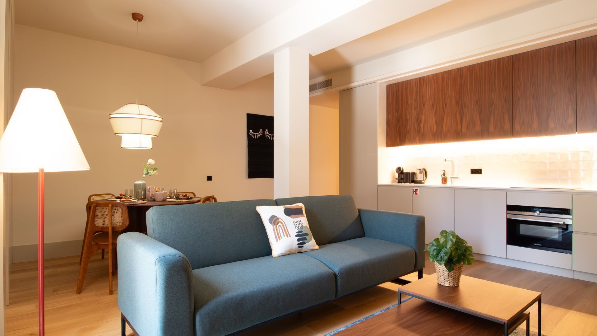 Ein modernes Wohnzimmer mit einer Couch, einem Esstisch mit Stühlen, einer Lampe und einer Küchenzeile mit Holzschränken und Geräten.