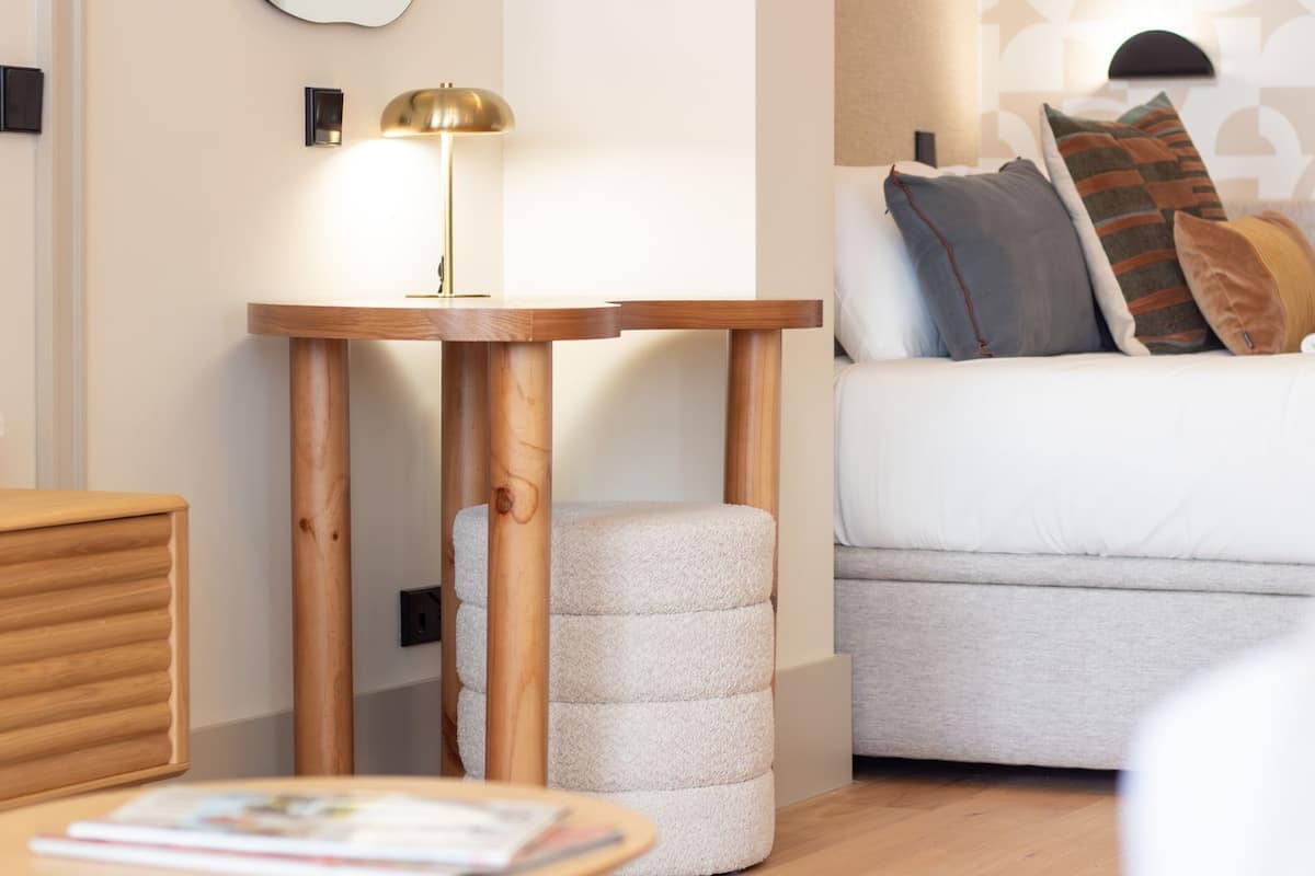 A modern bedroom with a wooden bedside table, a round lamp, a sofa with patterned cushions, and a bedside drawer.