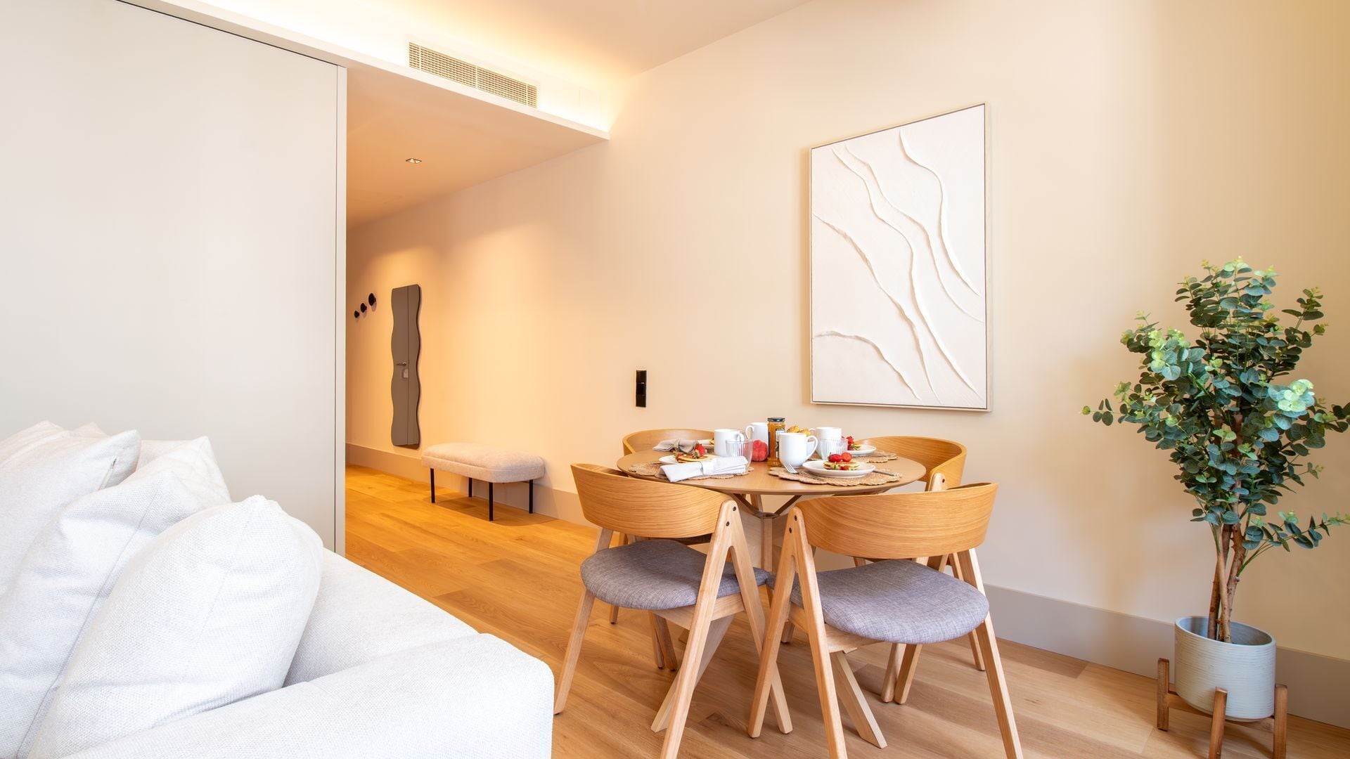 A modern hotel room with a wooden dining table set for breakfast, a bench, and a potted plant.