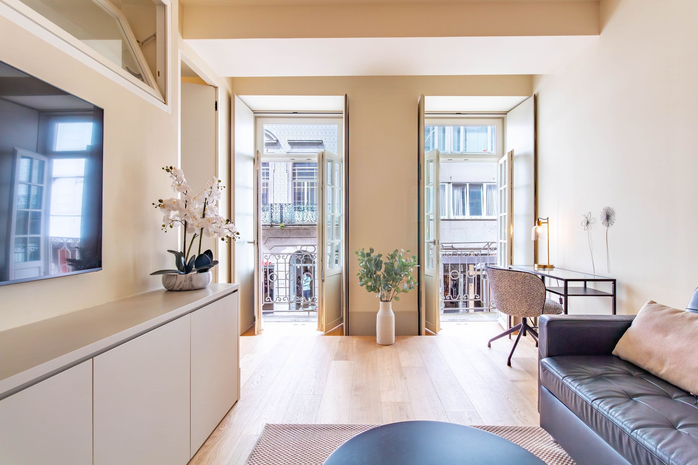 A modern living room with a sofa, a coffee table, and a TV on the wall. There are two doors leading to a balcony.
