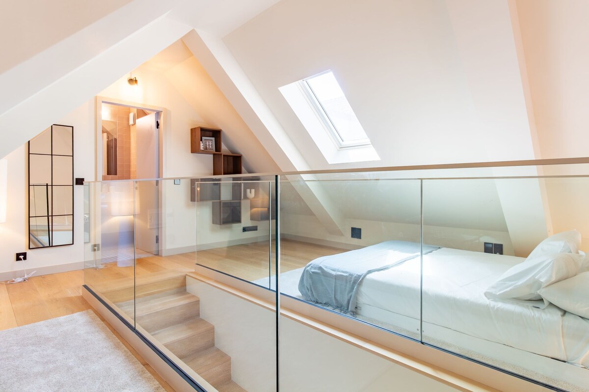 A modern bedroom with a glass railing, a bed, a staircase, and a skylight.