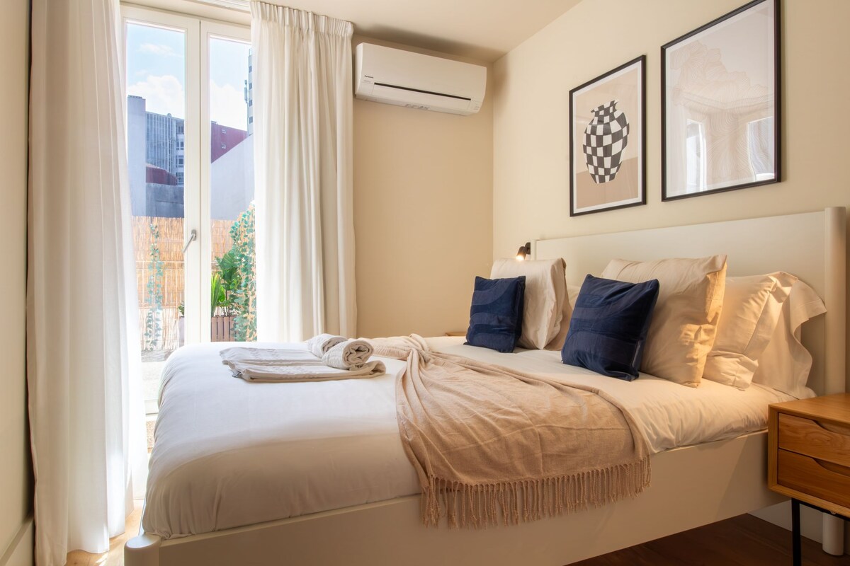 Habitación de hotel con cama, mesita de noche, ventana con cortinas, aire acondicionado y dos cuadros en la pared.