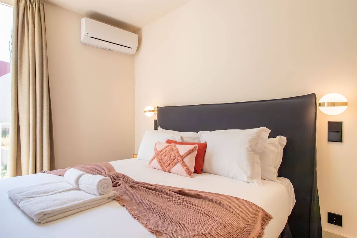 A hotel room with a bed, a window with curtains, an air conditioning unit, and a wall-mounted light fixture.