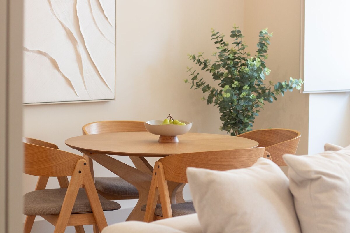 A dining area with a round wooden table, two wooden chairs, a bowl of fruit, a potted plant, and a white couch with pillows.