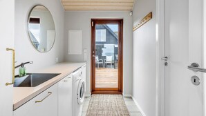 A narrow hallway with a wooden ceiling, a white sink with a gold faucet, a round mirror, a washing machine, and a door leading to an outdoor area with a view of a building.