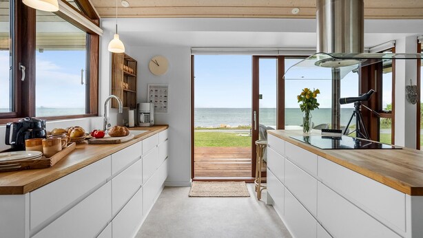 A modern kitchen with a view of the sea, featuring a wooden countertop, white cabinets, and a coffee machine.