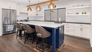 Cocina moderna con isla central, taburetes de bar, electrodomésticos de acero inoxidable, iluminación colgante y armarios blancos.
