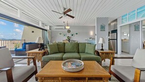 A spacious living area with a green sofa, wooden coffee table, and wicker chairs. A ceiling fan and a view of the ocean are visible. - The Caribbean Innisfree 1BR/2BA @Point Pleasant/Private Balcony/3 Pools/Beach! (Estate Smith Bay, East End Quarter)