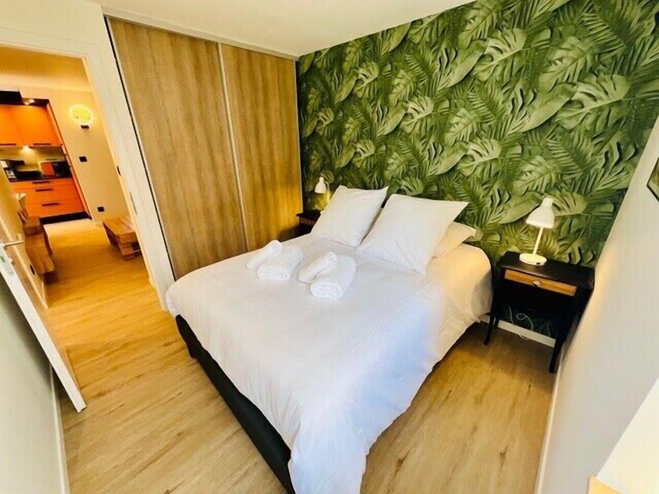 Quarto de hotel com uma cama, mesa de cabeceira, abajur e parede com padrão de folhas verdes.