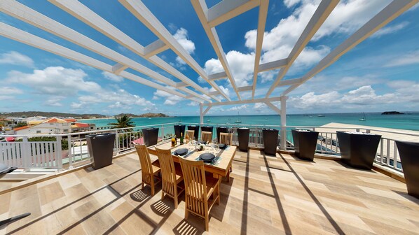 A terrace with a wooden table and chairs, a white pergola, and a view of the sea and buildings.