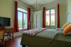 A bedroom with a bed, a desk with a TV, a chandelier, and a window with curtains.