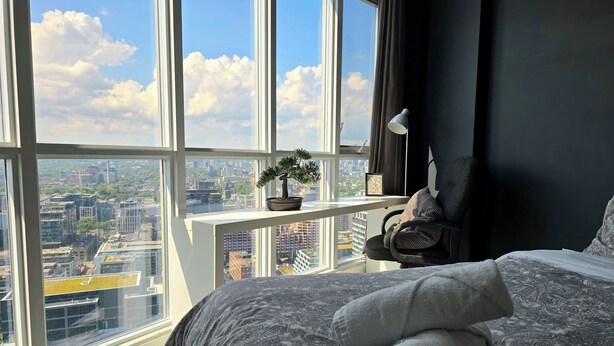 Ein Schlafzimmer mit großem Fenster, einer Topfpflanze, einem Sessel und einem ordentlich bezogenen Bett mit aufgerollten Handtüchern.