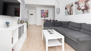A modern living room with a grey sofa, a white coffee table, and a flat-screen TV mounted on the wall. There are decorative items on a shelf and a painting of flowers on the wall. - Vivelorural Las Coquetas, Caleta de Fuste (Antigua)