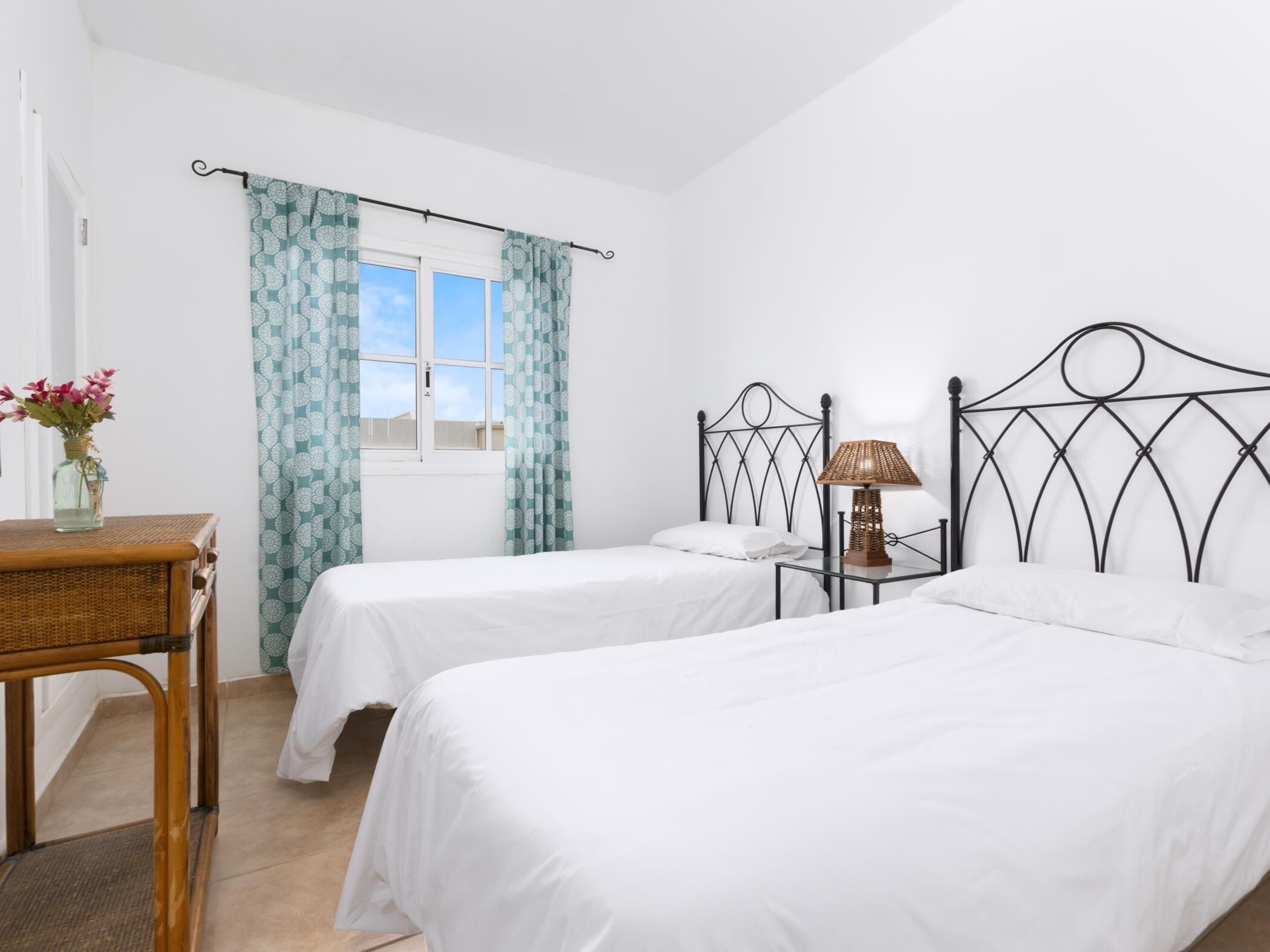 A bedroom with two single beds, a bedside table with a lamp, a wicker table with a vase of flowers, and a window with patterned curtains.