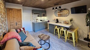 A compact living space with a kitchenette, a sofa with cushions, a dining area with stools, and a TV mounted on the wall. - Studio with balcony - near lake (Les Deux Alpes)