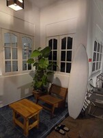 Una habitación con una mesa de madera y dos sillas, una planta grande en maceta y una tabla de surf apoyada contra la pared.