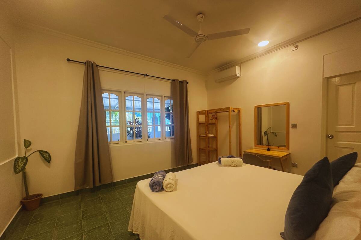 Ein Schlafzimmer mit einem Bett, einem Deckenventilator, einem Fenster mit Vorhängen, einem Holzschrank, einem Spiegel und einer Topfpflanze.