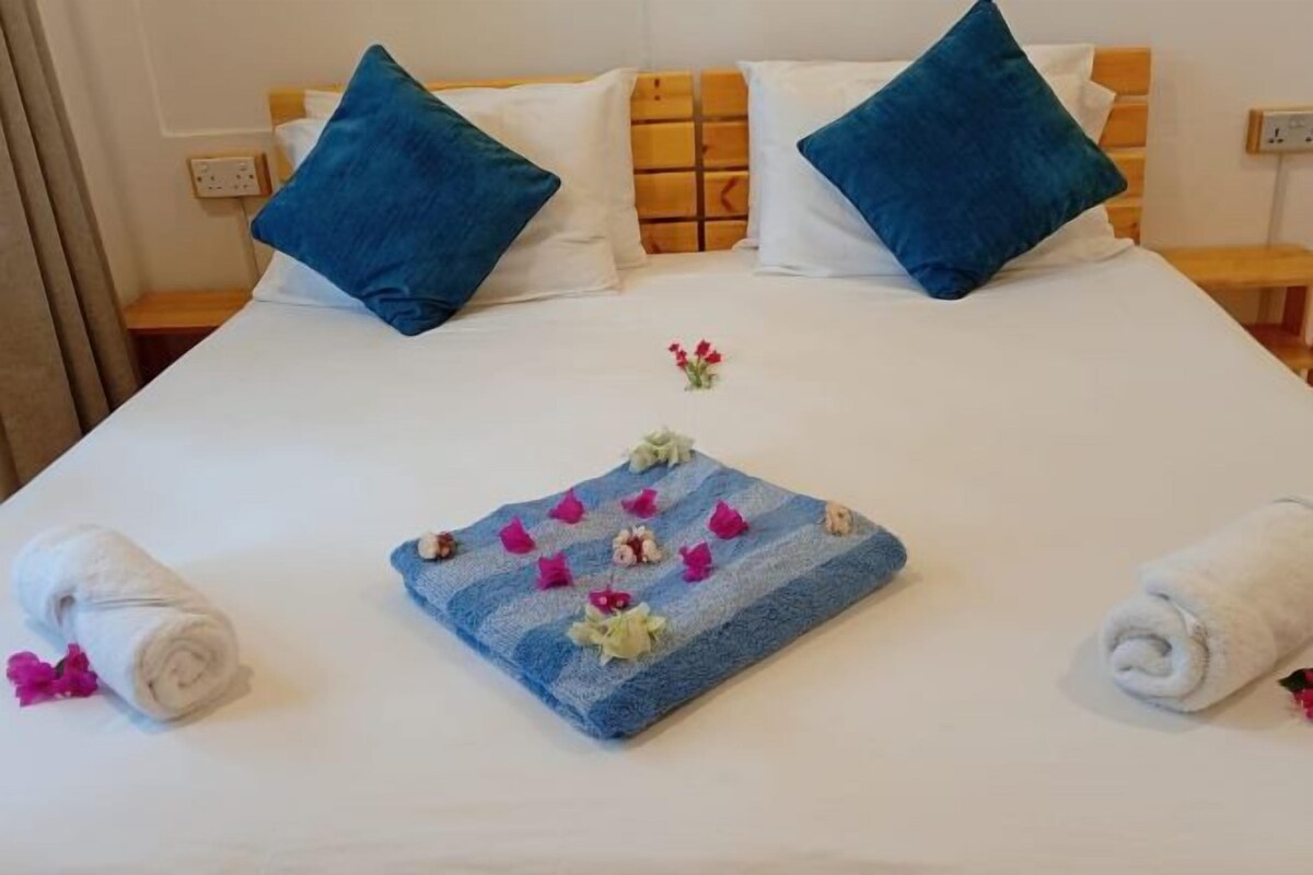 Een net opgemaakt bed met blauw-witte kussens, een blauwe handdoek met bloemen, en opgerolde handdoeken aan weerszijden.