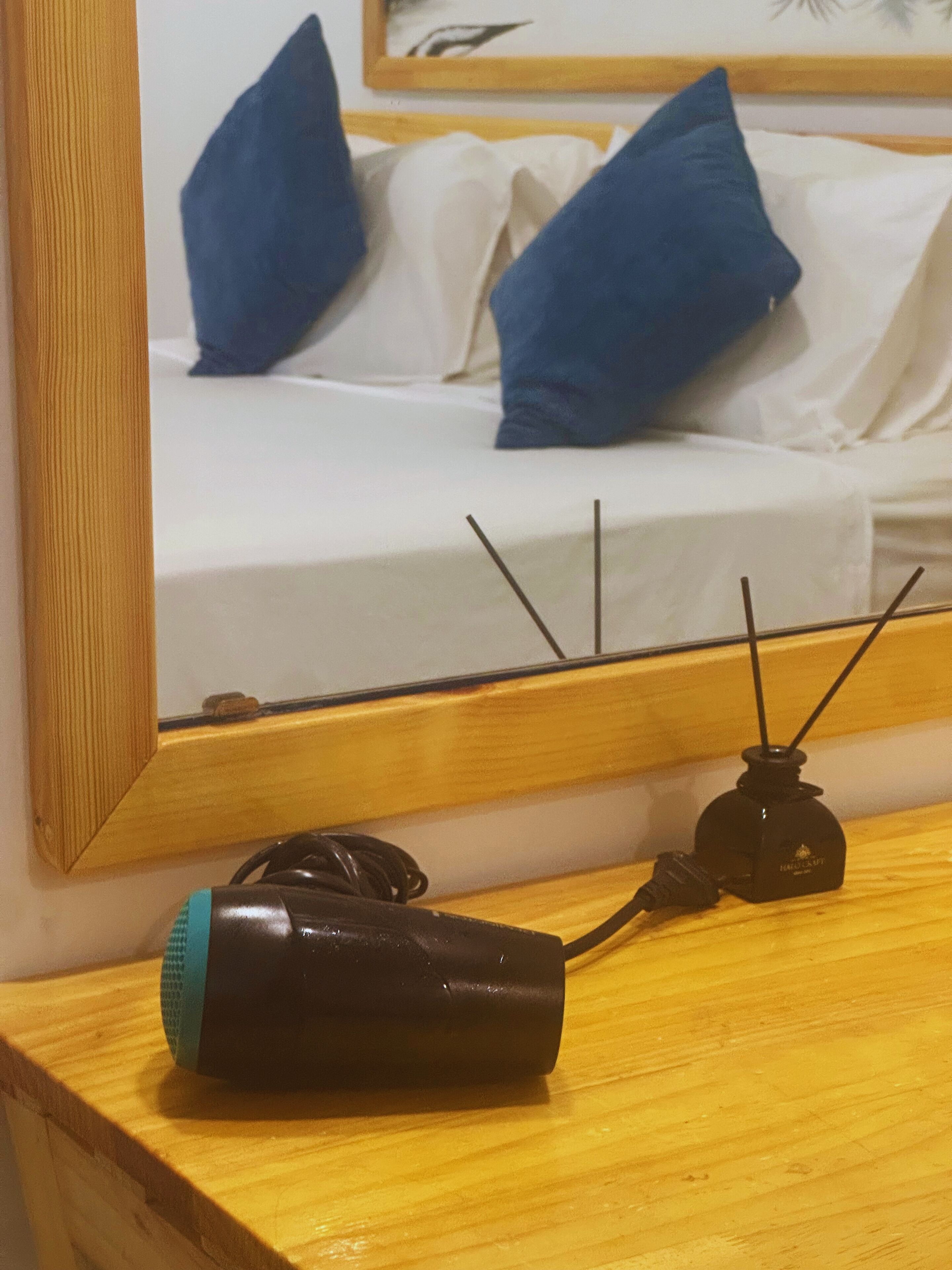 Una mesita de noche de madera con un dispositivo electrónico negro y un objeto azul, un espejo que refleja una cama con almohadas blancas y azules.