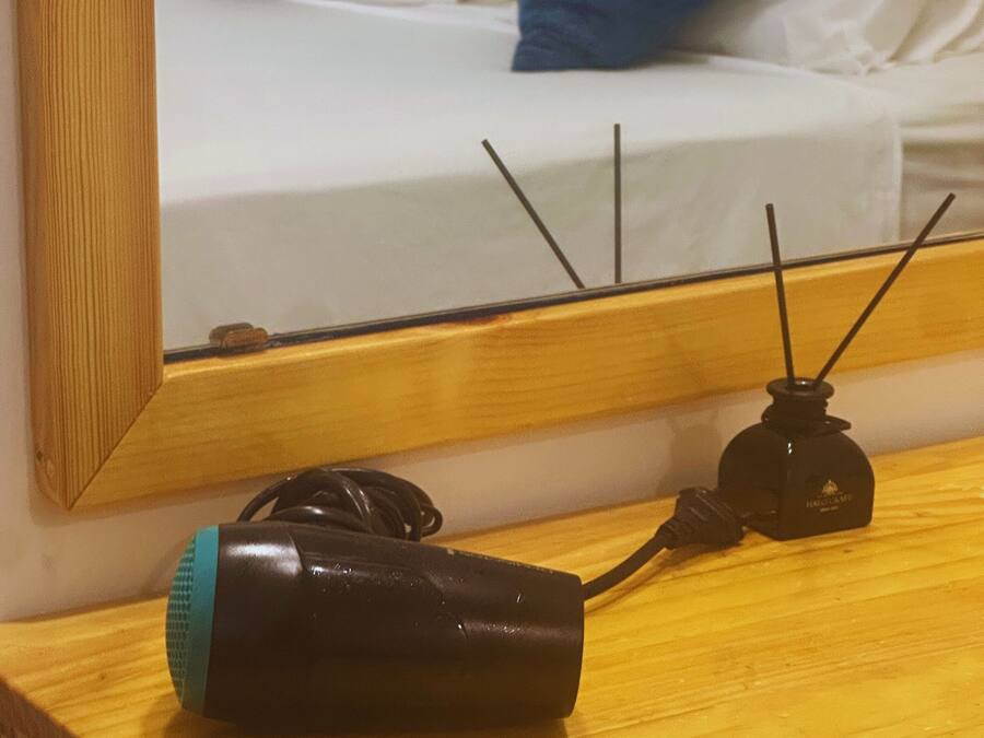 A wooden bedside table with a black electronic device and a blue object, a mirror reflecting a bed with white and blue pillows.