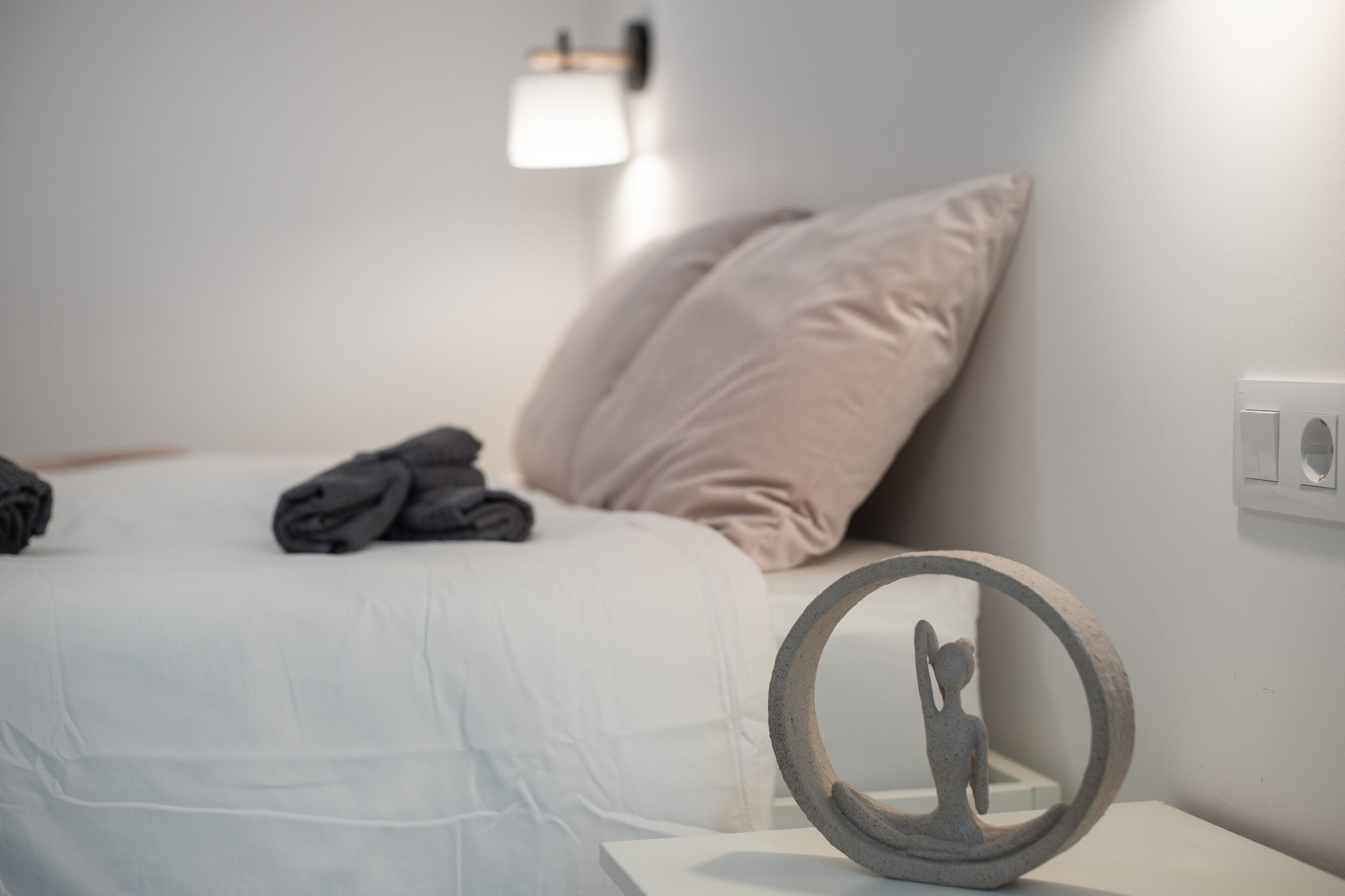 Una cama bien tendida con almohadas y toallas dobladas, una lámpara de pared y un objeto redondo sobre una mesita de noche.