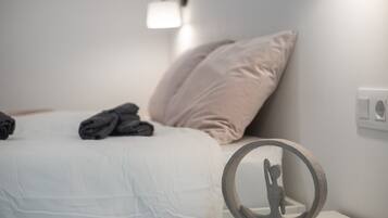 Una cama bien tendida con almohadas y toallas dobladas, una lámpara de pared y un objeto redondo sobre una mesita de noche.