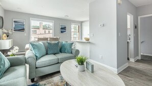 A modern living room with a grey sofa, a round coffee table, and a view of a building through large windows. - Lovely 1-bedroom apartment with Balcony, Wi-Fi, AC - only 30 mins to NYC (New Rochelle)
