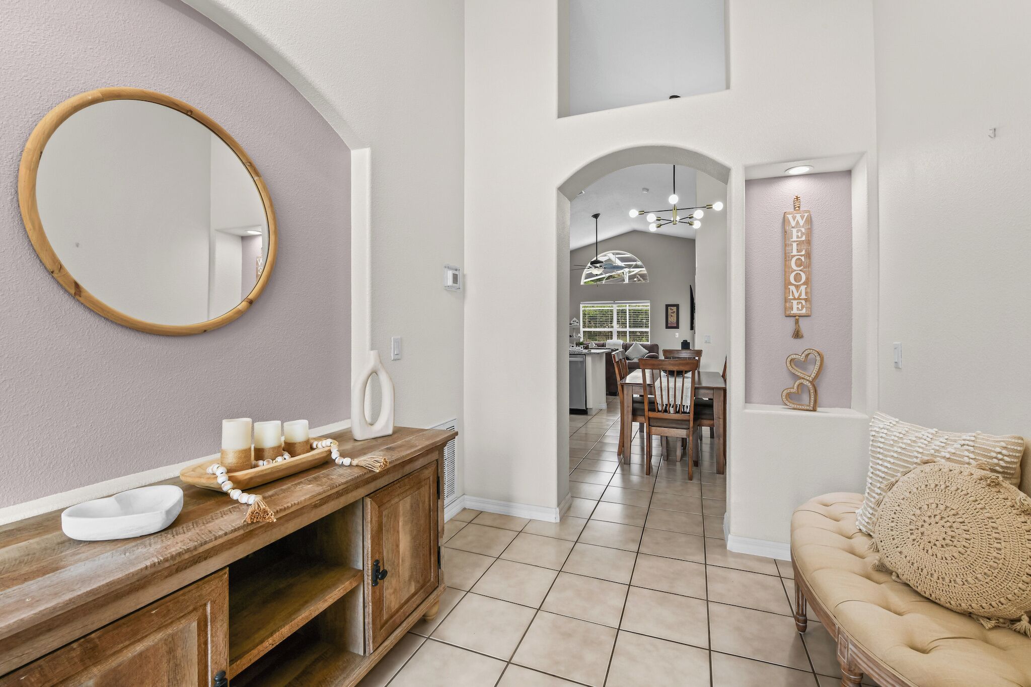 A modern interior with a wooden vanity, a round mirror, a beige sofa, and a dining area with a chandelier.
