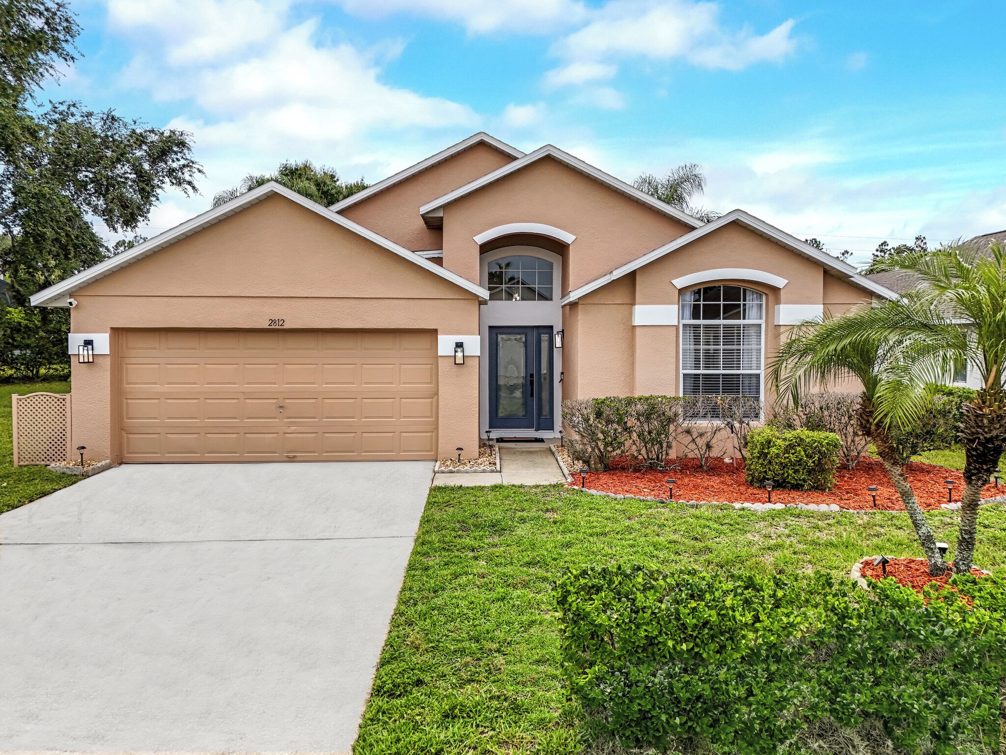 A two-story house with a beige exterior, a garage, and a well-maintained front yard with shrubs and a palm tree.