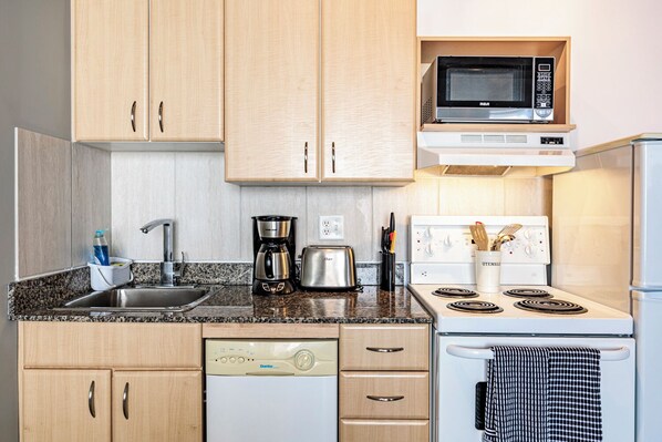A compact kitchen with wooden cabinets, a microwave, a coffee maker, a toaster, a dishwasher, and a stove. - Furnished to Impress—Steps from Everything! (Edmonton)