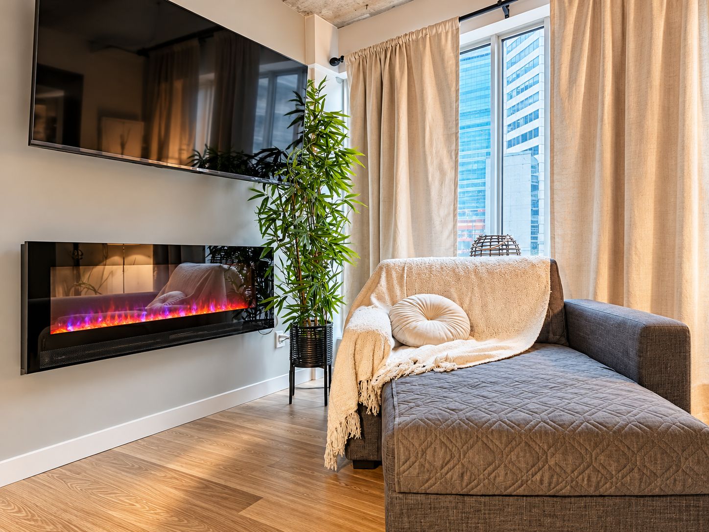 Un salon moderne avec un téléviseur à écran plat fixé au mur, un canapé agrémenté d’un plaid, une plante en pot et une fenêtre avec des rideaux.