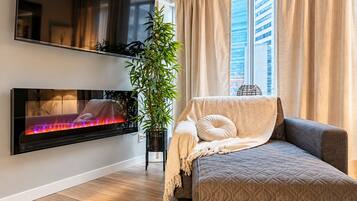 Un salon moderne avec un téléviseur à écran plat fixé au mur, un canapé agrémenté d’un plaid, une plante en pot et une fenêtre avec des rideaux.