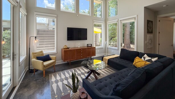 A spacious living room with large windows, a flat-screen TV, a sofa, a coffee table, and a cabinet. - New! The Mel~ Modern, Airy Cottage~5 Mins to Beach + UNCW (Wilmington)