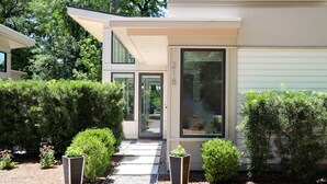A modern house with a covered patio, a large sliding glass door, and a well-maintained garden with various shrubs and a gravel pathway. - New! The Mel~ Modern, Airy Cottage~5 Mins to Beach + UNCW (Wilmington)
