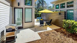 A modern house with a patio area featuring a table and chairs, a yellow umbrella, and a small barbecue grill. - New! The Mel~ Modern, Airy Cottage~5 Mins to Beach + UNCW (Wilmington)