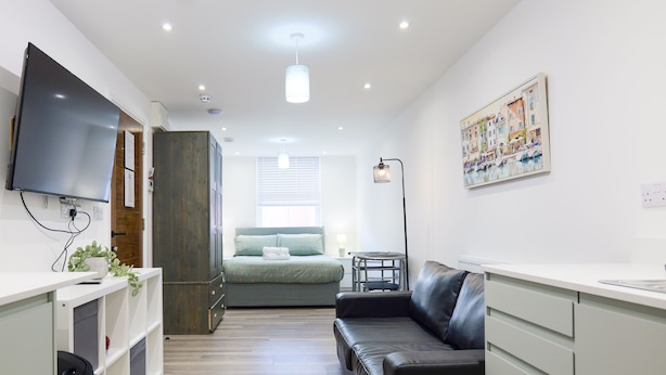 Una habitación moderna y bien iluminada con una televisión de pantalla plana, una cama con cubrecama verde, un sofá de cuero y una mesita de noche con una lámpara.