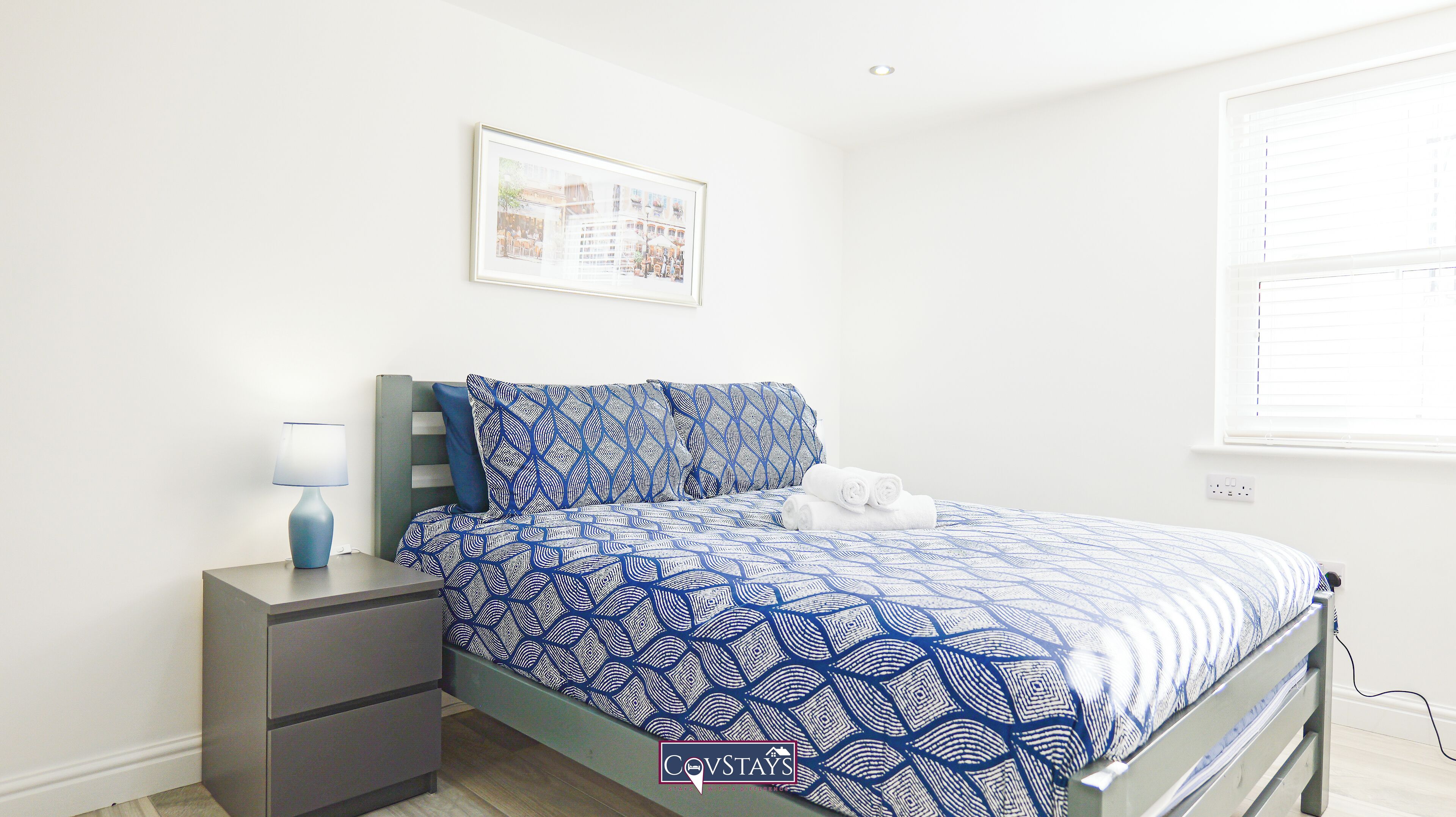 Un dormitorio con cama, mesita de noche, lámpara, un cuadro en la pared y ventana con persianas.