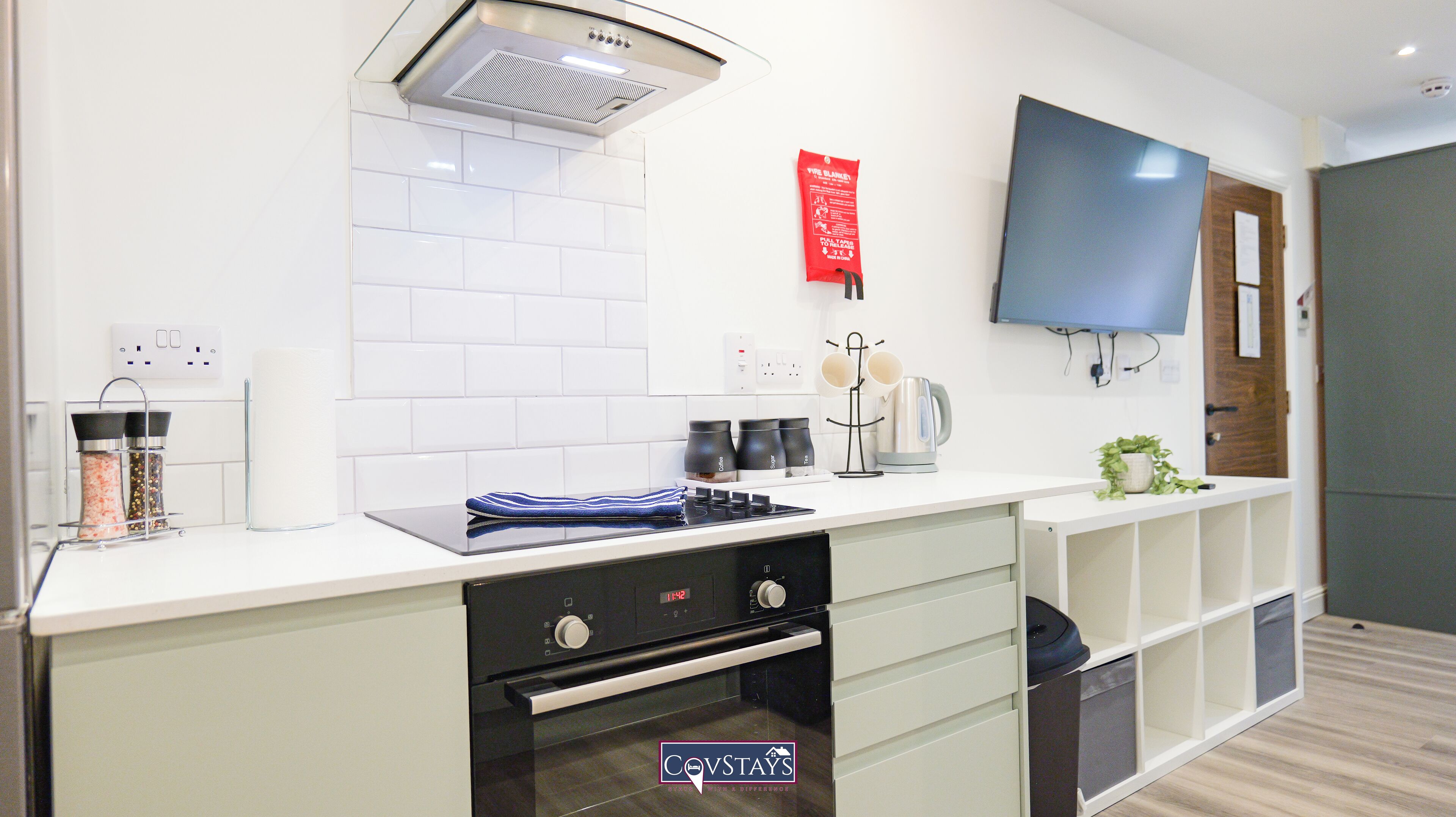 A modern kitchen with a built-in oven, a microwave, and a kettle on the counter. There is a TV mounted on the wall and a red fire blanket hanging on the wall.