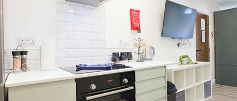 A modern kitchen with a built-in oven, a microwave, and a kettle on the counter. There is a TV mounted on the wall and a red fire blanket hanging on the wall.