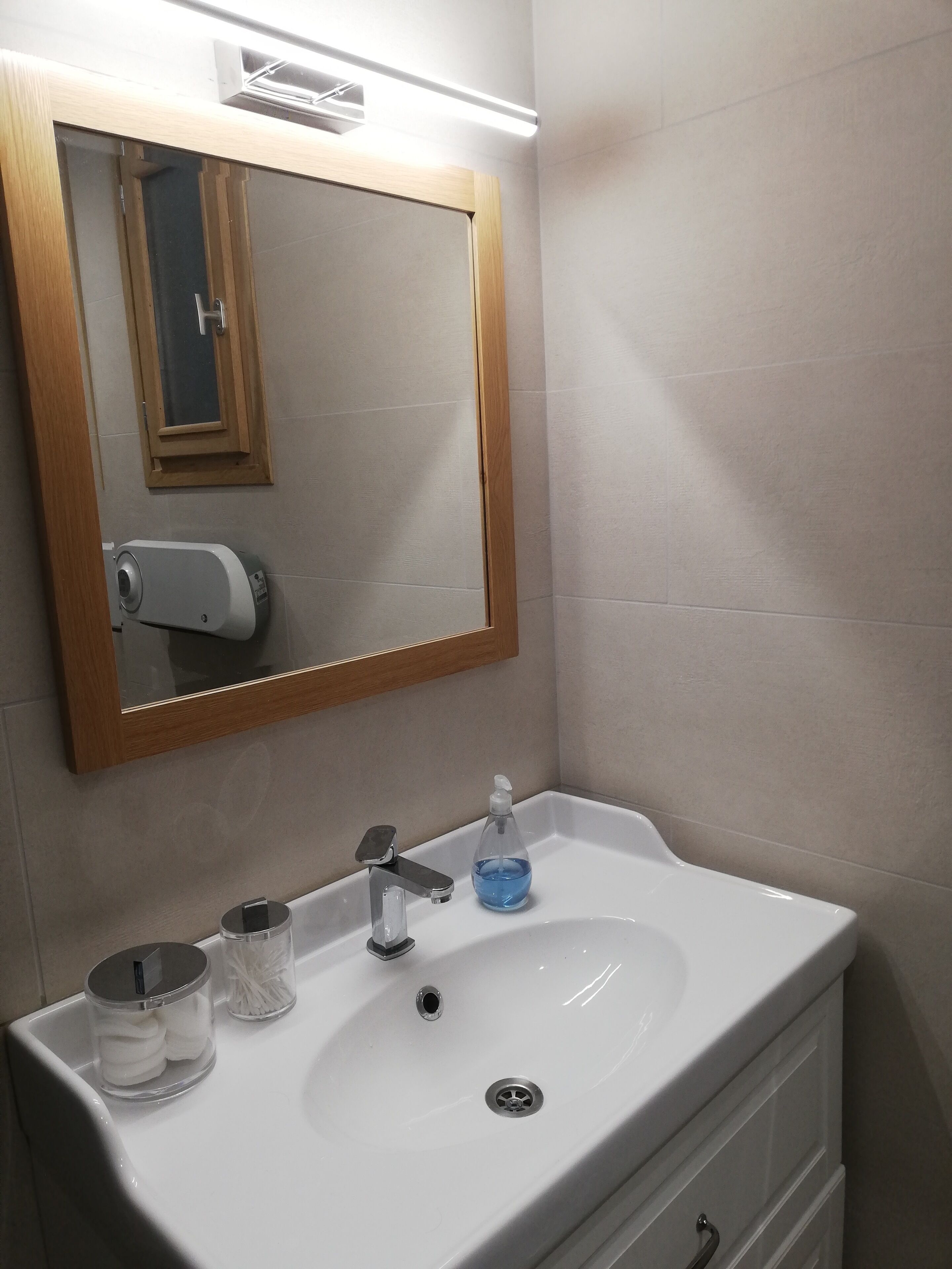 A bathroom with a white sink, a mirror with a wooden frame, a soap dispenser, and a tissue holder.