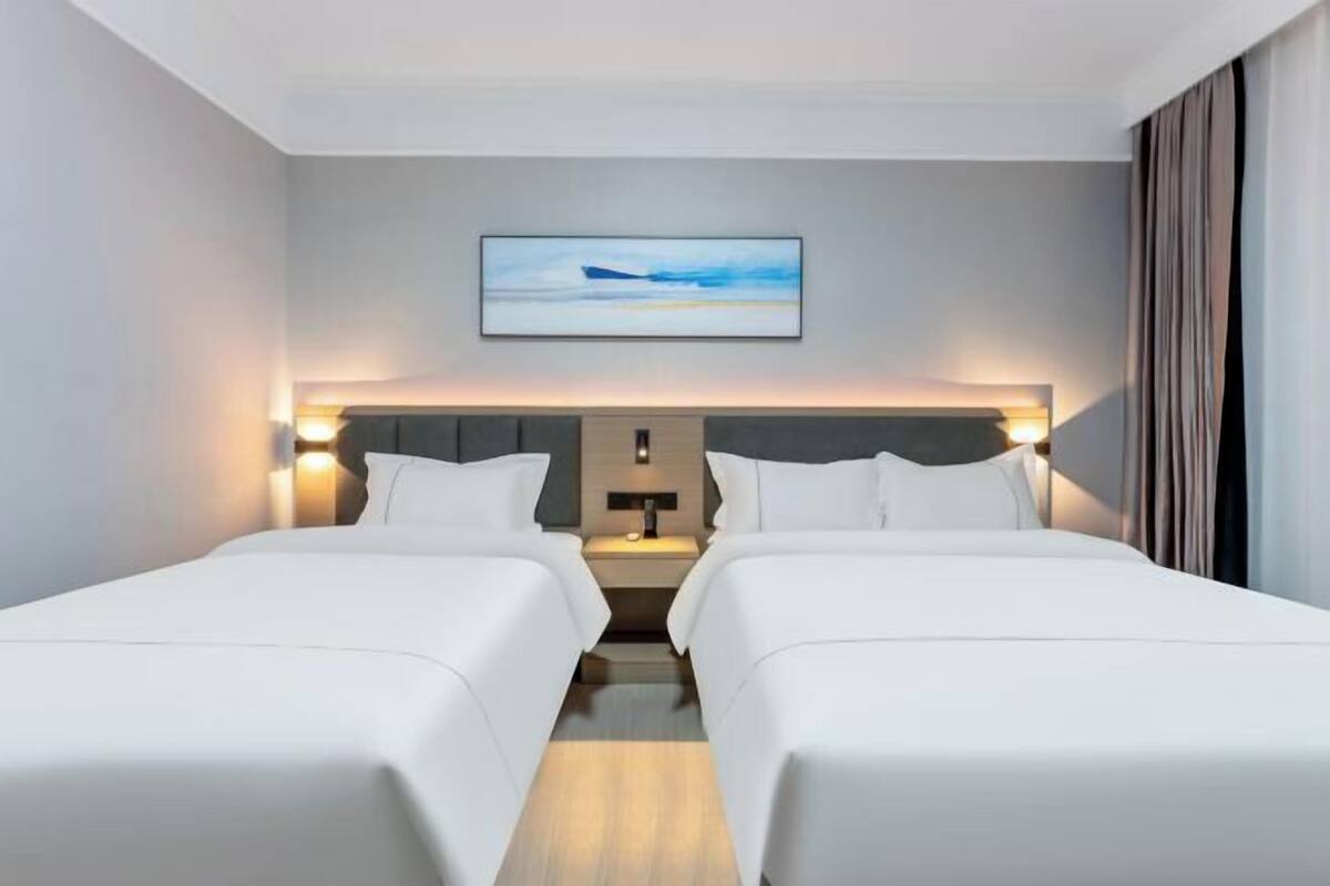 A hotel room with two beds, a nightstand, a wall-mounted artwork, and a window with curtains.