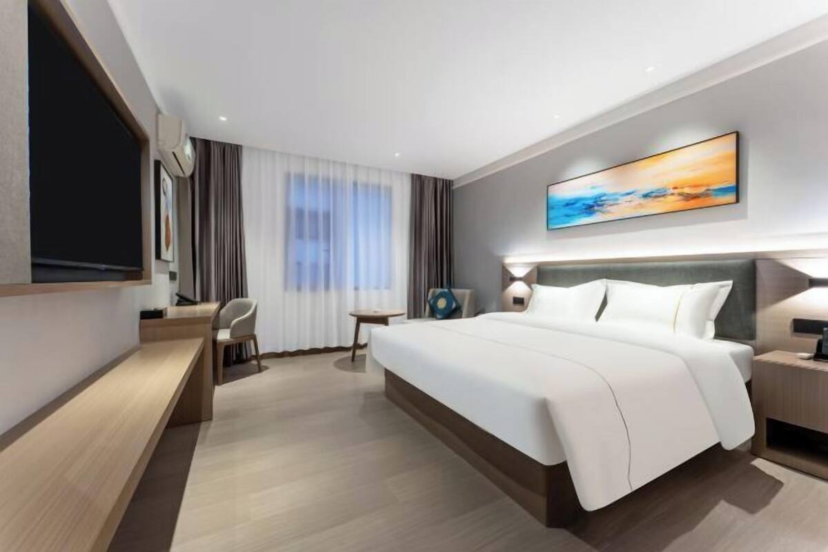 A modern hotel room with a large bed, a desk, a chair, a TV, and a painting on the wall.