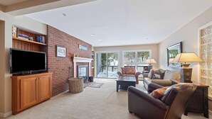A living room with a brick fireplace, a flat-screen TV, a sofa, and a coffee table. - Ski-In/Out at Okemo | 3BR Ledgewood w/ Fireplace (Ludlow)