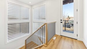 A modern interior with a wooden staircase, white walls, and large windows with horizontal blinds.
