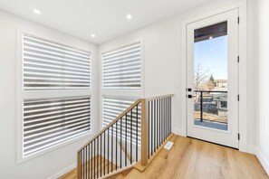 A modern interior with a wooden staircase, white walls, and large windows with horizontal blinds.