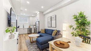A modern living room with a blue sofa, a dining table with a wooden board, and a kitchen in the background.