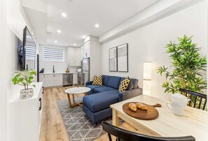 A modern living room with a blue sofa, a dining table with a wooden board, and a kitchen in the background.