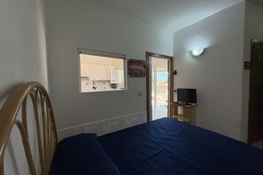 A bedroom with a bed, a wooden chair, a television on a stand, and a window overlooking a kitchen.