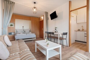 Ein modernes Wohnzimmer mit einer Couch, einem Esstisch mit Stühlen, einer Küchenzeile und einem an der Wand montierten Fernseher.